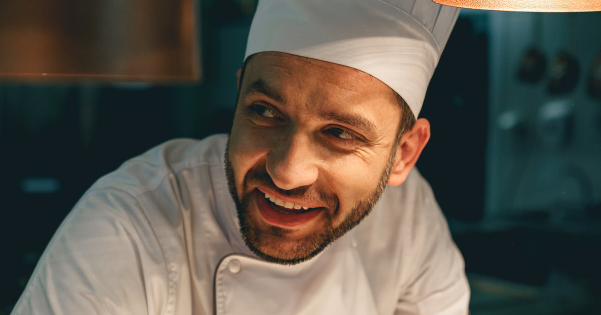 Close up of smiling male chef in uniform standing on kitchen of restaurant and looking away