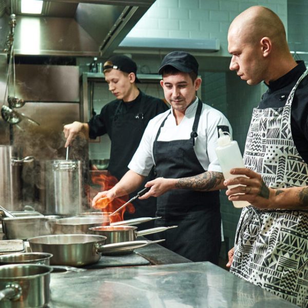 Dangerous work. Profesional chef teaching his two young trainees how how to flambe food safely. Cooking techniques