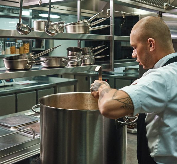 Focused on his work. Side view of famous young chef with tattoos on his arms cooking a soup in a restaurant kitchen
