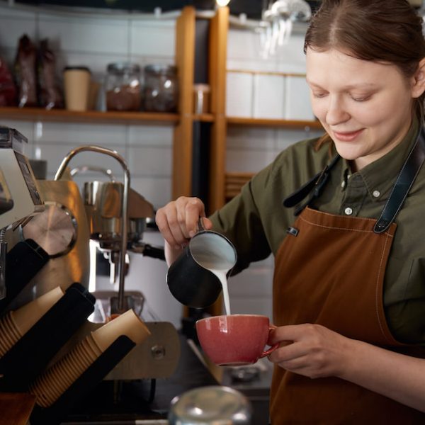 Experienced woman barista pouring milk into coffee cup with freshly brewed drink