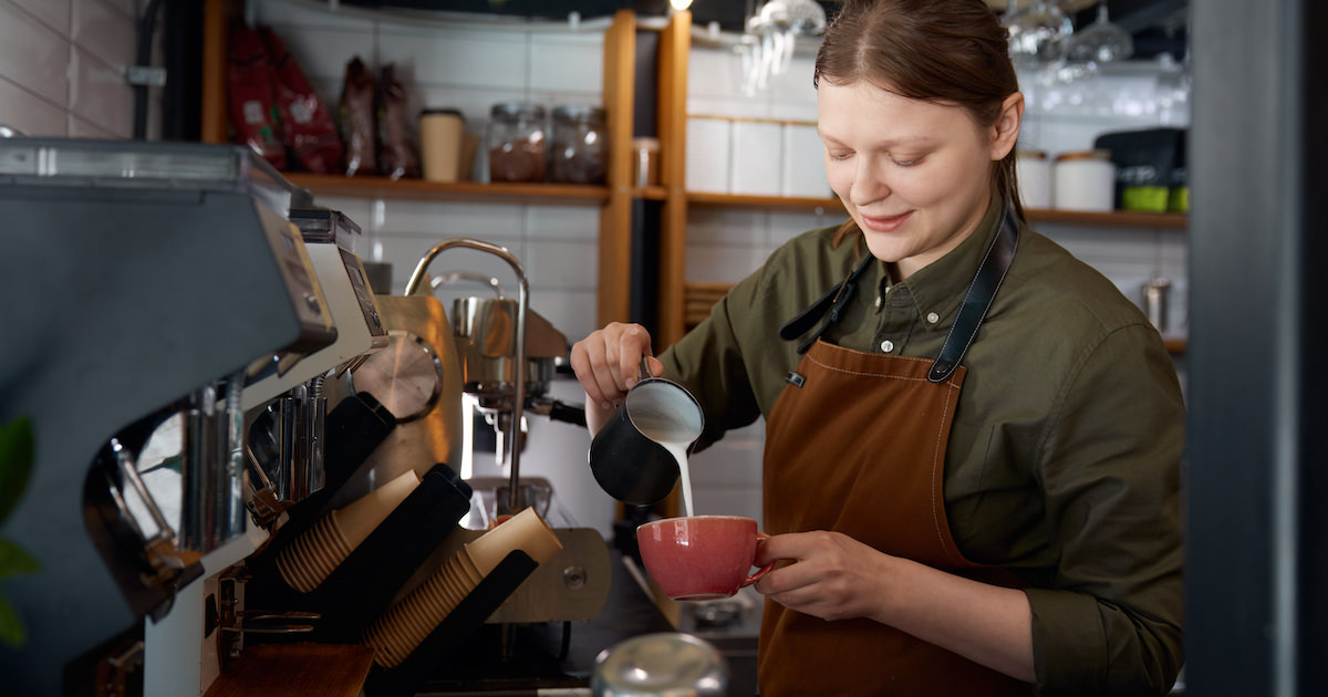 Experienced woman barista pouring milk into coffee cup with freshly brewed drink
