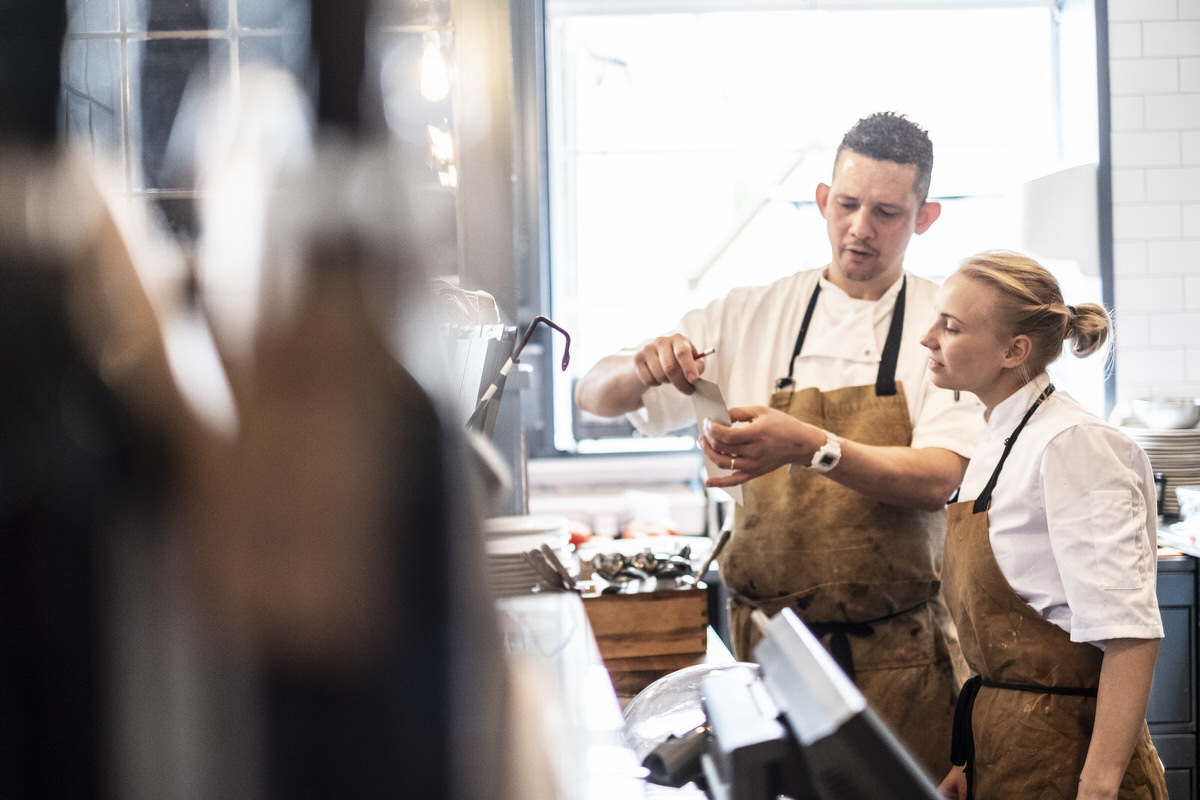 Male and female chef wearing brown aprons standing at a counter, checking an order.