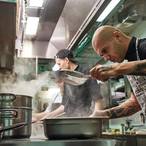 Restaurant kitchen. Professional chef in apron, with tattoos on his arms is sifting the flour while cooking with his two assistants
