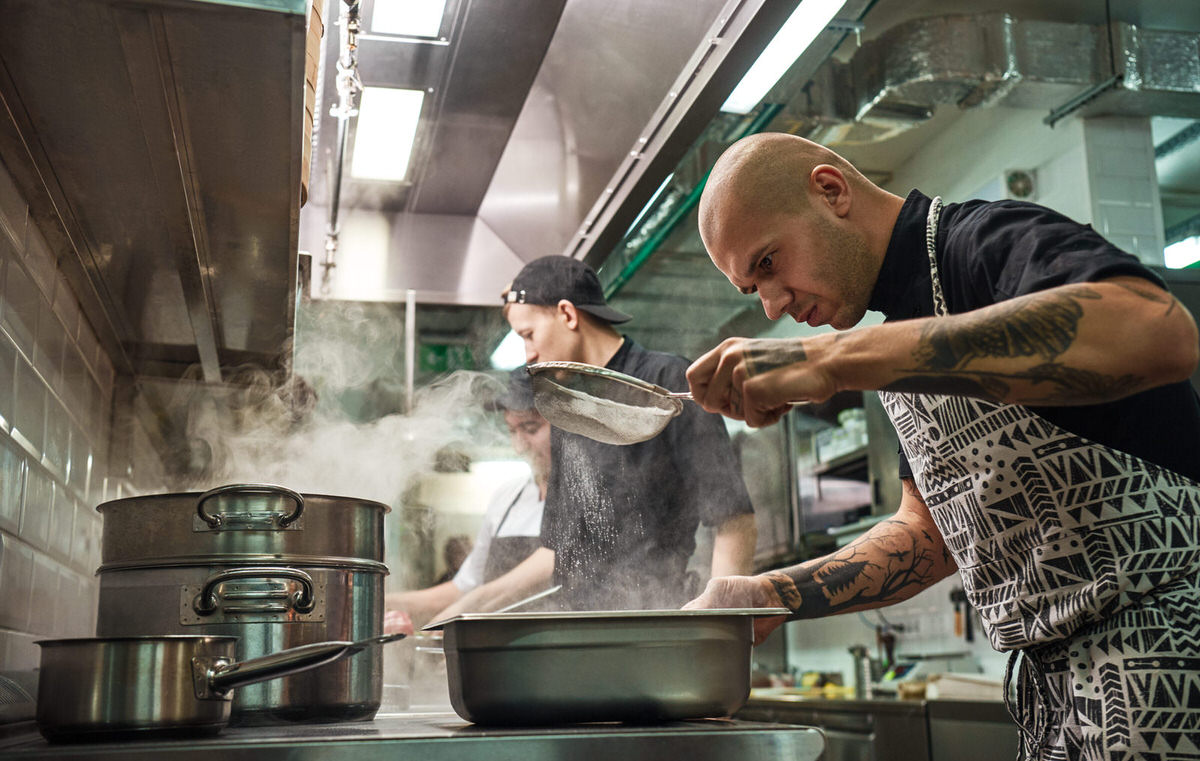 Restaurant kitchen. Professional chef in apron, with tattoos on his arms is sifting the flour while cooking with his two assistants