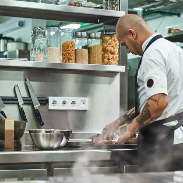 Working process. Back view of male chef in apron and with tattoos on his arms cutting a meat while standing in a restaurant kitchen.